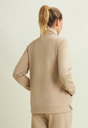 Donna con coda di cavallo bionda, che indossa una maglia a maniche lunghe beige e pantaloni abbinati, in piedi con le mani in tasca, voltata di spalle rispetto alla fotocamera.