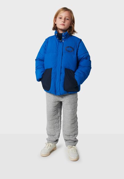 Blaues Pufferjackett mit marineblauen Akzenten und Taschen, ausgestattet mit einem hohen Kragen. Graue gesteppte Hose und weiße Sneaker vervollständigen das Outfit.