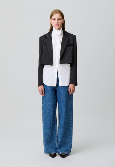 Blazer negro corto sobre una camisa blanca, combinado con jeans de mezclilla azul de pierna ancha. El blazer presenta hombros estructurados y un bolsillo lateral.