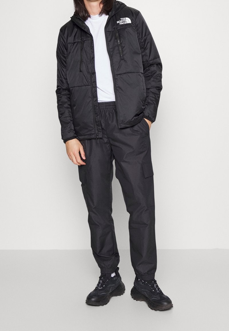 Personne portant une doudoune noire North Face, un pantalon cargo noir, des baskets noires et un t-shirt blanc, debout devant un fond uni.