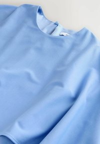 Šviesiai mėlyna bluzelė, lygaus paviršiaus, apvaliu iškirpimu, trumpomis rankovėmis, sagos uždarymu nugarinėje pusėje, minimalistinio dizaino be matomų raštų.