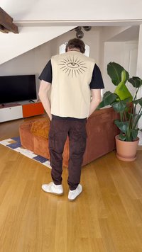 Beige Weste mit schwarzer Augen-Grafik auf dem Rücken, kombiniert mit dunkelbraunen Cargo-Hosen und weißen Turnschuhen. Holzboden und Pflanzen im Hintergrund.