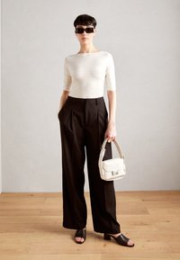 Cremefarbener, taillierter Oberteil, hochgeschnittene schwarze Plisseehose, dunkle Sandalen und eine kleine weiße Handtasche mit einem Riemen, vor einem neutralen Hintergrund.