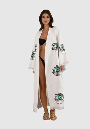 Frau trägt einen langen weißen offenen Bademantel mit grünen und schwarzen Augenmustern über einem schwarzen Bikini und gewebten Sandalen, steht vor einem schlichten Hintergrund.