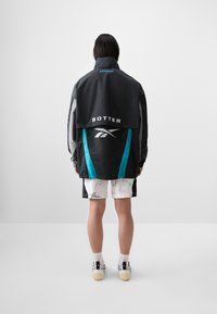 Black and gray oversized jacket with turquoise accents, featuring "BOTTTER" logo and Reebok symbol. Worn over white shorts with signatures.