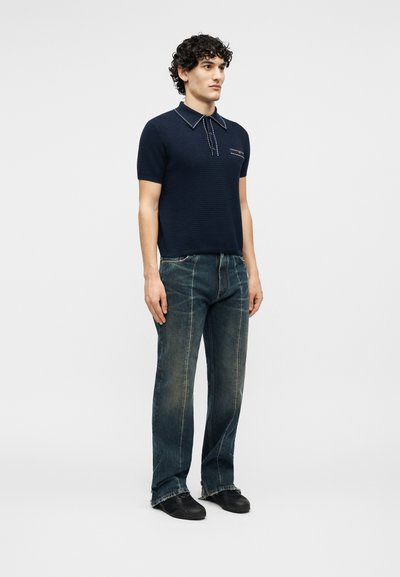 Άνδρας με σγουρά μαλλιά στέκεται φορώντας μπλε ναυτικό πλεκτό πόλο με κοντά μανίκια, ξεβαμμένα μπλε τζιν με ακατέργαστες άκρες και μαύρα παπούτσια σε μονόχρωμο φόντο.