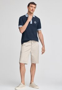 Marinblå polo skjorta med ljusblå krage och manschetter, kombinerad med beiga shorts och ljusa sneakers. Har en broderad logotyp på bröstet.