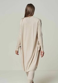 Helle beige lange Strickjacke mit seitlichen Taschen, gerippter Textur, offener Vorderseite und abgerundetem Saum. Die Rückansicht zeigt ein einfaches, lässiges Design.