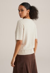 Béžový pletený sveter s textúrovaným vzorom, krátkymi rukávmi a priliehavým lemom. Nosený cez hnedú sukňu, pohľad zozadu.