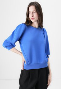 Felpa blu con maniche a sbuffo, scollatura rotonda e orlo a costine. Tessuto liscio e design aderente, abbinata a pantaloni neri.