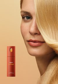 Spray leave-in condizionante Dyson Omega in una bottiglia rosso corallo con beccuccio giallo, esposto accanto a una modella con capelli lisci e biondi.