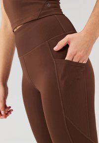 Leggings marrones de cintura alta en tejido suave con un bolsillo lateral, que presentan textura acanalada en la banda superior y detalles de costura.