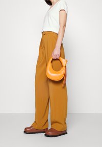 Gul-orange handväska med rundad form och en knutdetalj vid handtaget. Bärs med en ljusgrå t-shirt, bruna vida byxor och kraftiga skor.