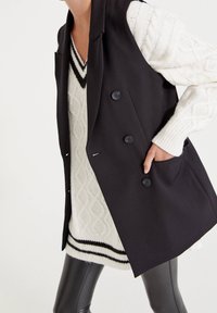 Czarna kamizelka o dopasowanym kroju z pięcioma guzikami i bocznymi kieszeniami, noszona na kremowym swetrze w sploty kabelkowe z czarnym paskiem na dole.