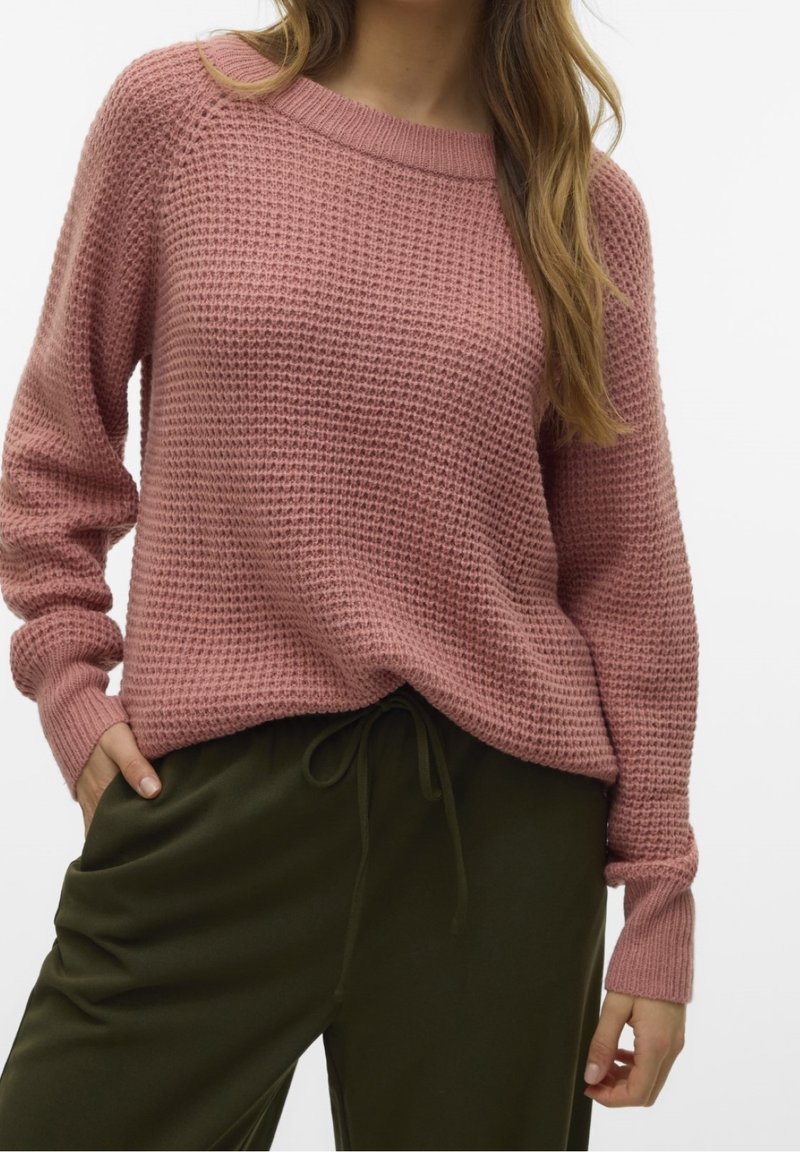 Femme portant un pull en maille texturé rose poussière associé à un pantalon à cordon vert olive foncé, les mains dans les poches, sur un fond uni.