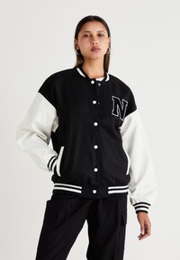 Veste varsity noire et blanche avec un grand patch en "N", des poignets et un ourlet côtelés, et des poches latérales, dotée d'un design à fermeture à boutons.