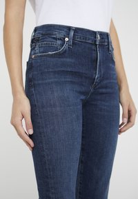 Mörkblå denimjeans har hög midja, framfickor och en figursydd form. Texturen är slät med subtila blekningar och sömnadsdetaljer.