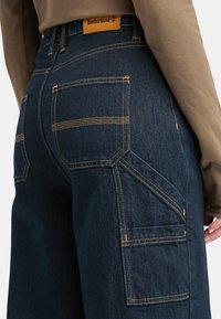 Jeans en denim bleu foncé avec des surpiqûres orange contrastantes, comportant plusieurs poches et une étiquette Timberland sur la ceinture.