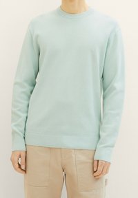 Person, der einen hellblauen strukturierten Langarmpullover und beigefarbene Hosen mit vorderen Aufsetztaschen trägt, steht vor einem schlichten Hintergrund.