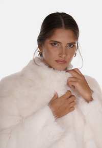 Cappotto in pelliccia sintetica bianca con colletto alto, morbida texture e maniche lunghe. La modella tiene il colletto, mettendo in mostra il design del capo.