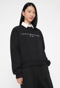 Juodas sweatshirt, pagamintas iš minkštos medžiagos, su baltu logotipu, kuriame yra užrašai "TOMMY HILFIGER" ir "EST. 1985", su gumuotu apvadu ir rankogaliais.