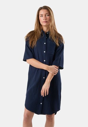 Młoda kobieta o długich blond włosach, ubrana w luźną granatową sukienkę zapinaną na guziki z krótkimi rękawami, stojąca z założonymi na siebie ramionami.