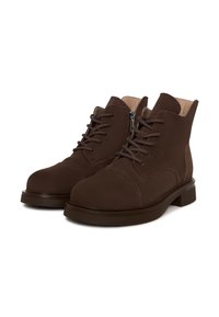 Botas de ante marrón hasta el tobillo con cordones, puntera redonda, superficie texturizada y cremallera lateral. Equipadas con una suela gruesa y oscura.