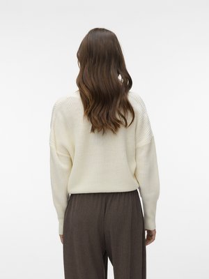 Ženska z dolgimi rjavimi lasmi, oblečena v kremasto pleten pulover in široke rjave hlače, stoji z obrnjenim hrbtom proti enostavni ozadju.