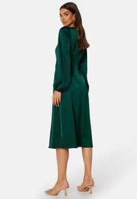 Zielona satynowa sukienka midi z długimi bufkami, dopasowaną górą, rozkloszowanym dołem i gładką fakturą. Przezroczyste sandały na obcasie uzupełniają całość.