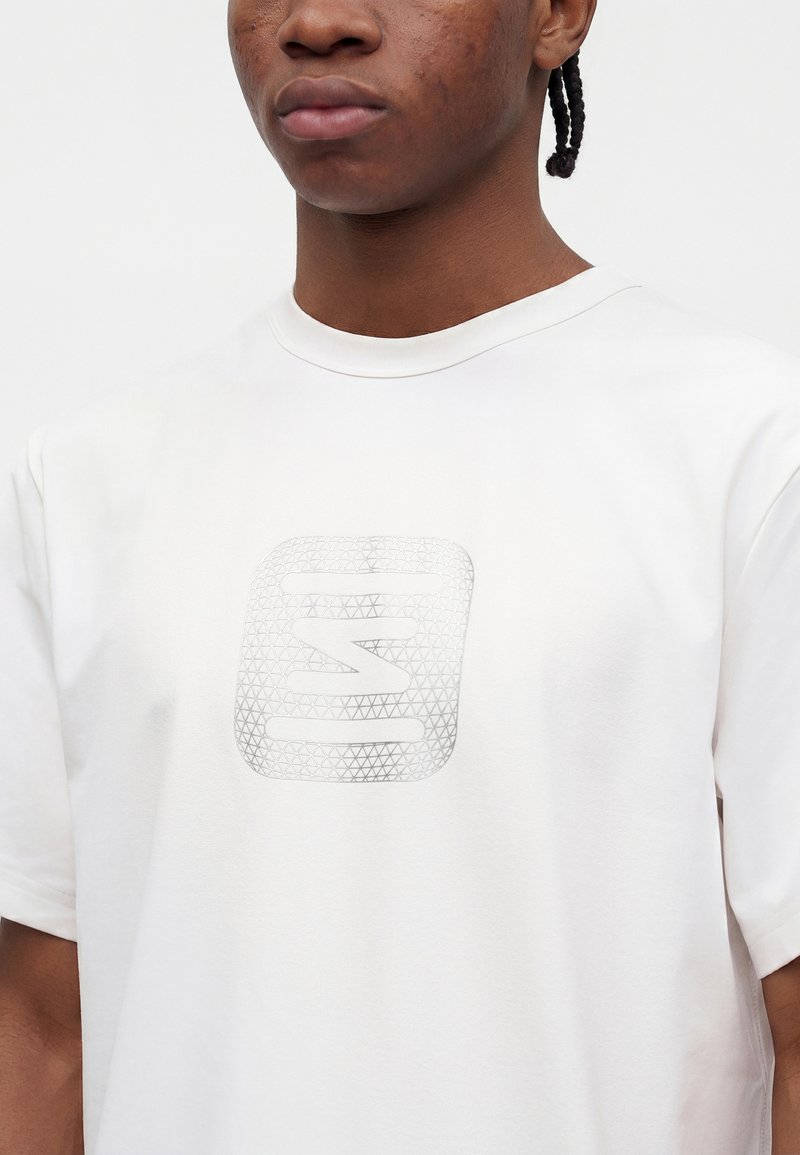 Young person wearing white T-shirt with a light gray geometric square logo featuring stylized letters centered on the chest.