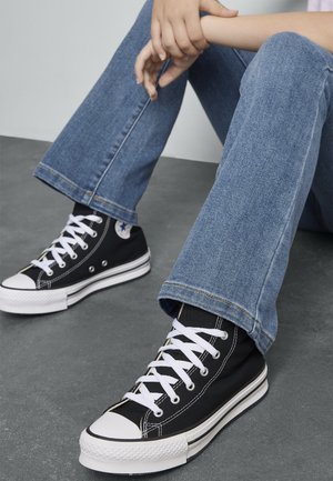 Mustat kangaskorkeavartiset tennarit valkoisilla kumipohjilla, joissa on valkoiset nauhat ja pyöreä logo; yhdistetty sinisiin levenevän lahkeen farkkuihin.