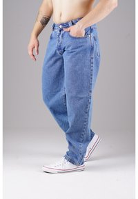 Helle blaue Denim-Jeans mit lockerer Passform, ausgestattet mit einem Reißverschluss und Knopfverschluss, vorderen Taschen und Gesäßtaschen. Getragen mit weißen Sneakern.