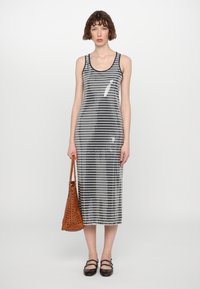 Robe débardeur pailletée avec des rayures verticales noires et argentées, mi-longue, décolleté rond. Modèle tenant un sac fourre-tout perforé marron.