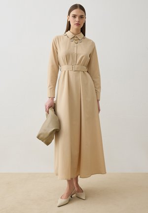 Ung kvinde iført en beige lang kjole med bælte og dekorativ krave, der holder en matchende taske, står på beige gulv mod en hvid væg.