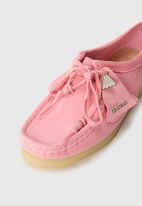 Pantof de suede roz, tip slip-on, cu vârf rotund, având un design cu șireturi, mărgele din lemn și o etichetă metalică. Talpa este de culoare bej deschis.