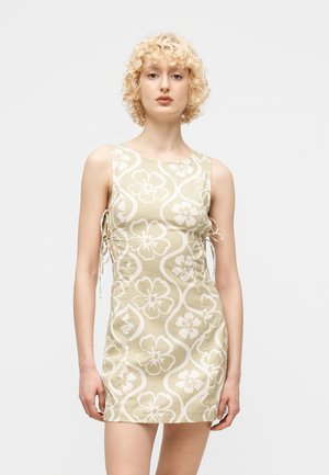 Νεαρό άτομο με κοντά  σγουρά ξανθά μαλλιά, φορώντας ένα μπεζ και άσπρο φλοράλ αμάνικο φόρεμα με δεσίματα στο πλάι, στέκεται μπροστά σε ένα απλό φόντο.