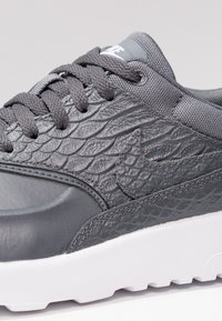 Baskets Nike grises avec un motif texturé en écailles sur la tige, des lacets gris, un col rembourré et une semelle blanche.