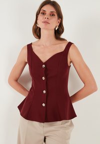 Burgundska peplum bluzica s dubokim V-izrezom, širokim naramenicama i pet sjajnih gumba sprijeda; glatka tkanina s strukturiranim krojem.