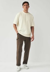 T-shirt oversize beige chiaro con scollatura rotonda, abbinato a pantaloni cargo aderenti marroni scuri con tasche laterali e orli risvolti. Sneakers bianche.