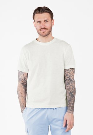 Hellbeige Kurzarm-T-Shirt aus weichem Stoff, mit Rundhals-Ausschnitt und lockerer Passform. Auffällige Tattoos sind am Arm sichtbar.