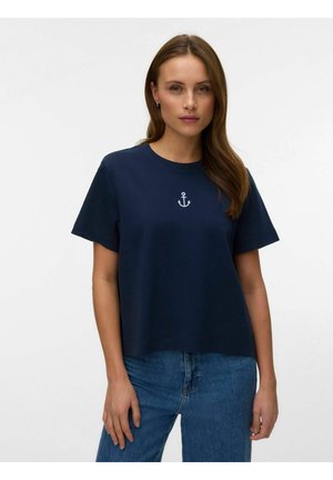 Kobieta w granatowym, krótkim rękawie koszulce z białym emblematem kotwicy na środku klatki piersiowej oraz niebieskich dżinsach na jednolitym tle.