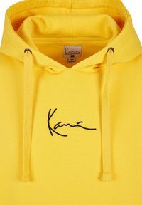 Sweat à capuche jaune avec cordons réglables et logo « Kani » en cursive noire brodé sur la poitrine, étiquette taille large visible à l'intérieur du col.