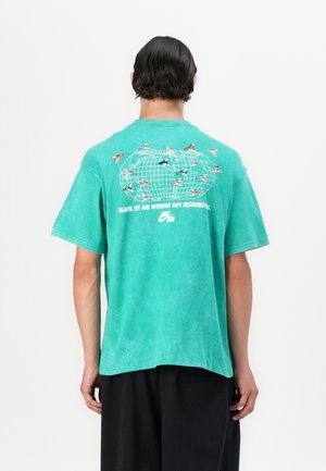Camiseta de algodón verde con un corte relajado, que presenta un gráfico de un globo terráqueo y unos zapatos en la parte posterior, junto con el texto "Viaja por aire sin reservas".