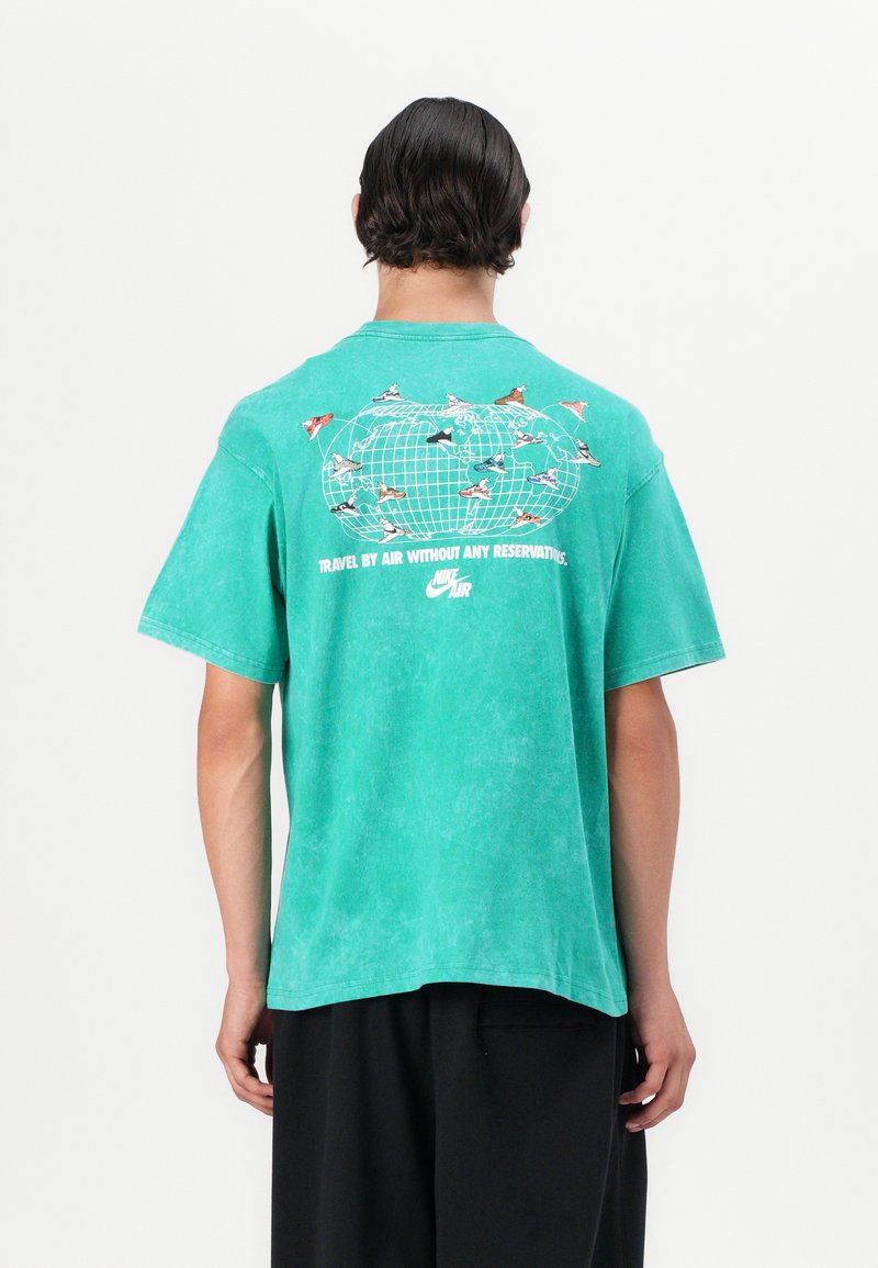 T-shirt de algodão verde com um corte descontraído, apresentando um gráfico de um globo e sapatos nas costas, juntamente com o texto "Viaje de avião sem reservas."