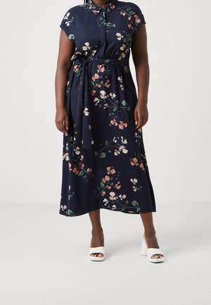 Vrouw draagt een marineblauwe midi-jurk met roze en witte bloemenpatronen, korte mouwen, een gestrikte tailleband en witte open-toe blokhakken.