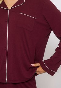 Pigiama sopra bordeaux con piping bianco, texture a coste, chiusura con bottoni e taschino sul petto. Maniche lunghe e design dalla vestibilità rilassata.