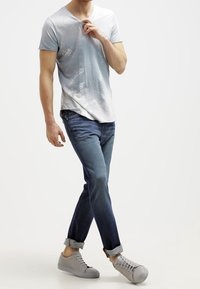 Camiseta de cuello en V azul claro con un patrón degradado y moteado, combinada con vaqueros azul oscuro y zapatillas grises, con el dobladillo enrollado en el denim.