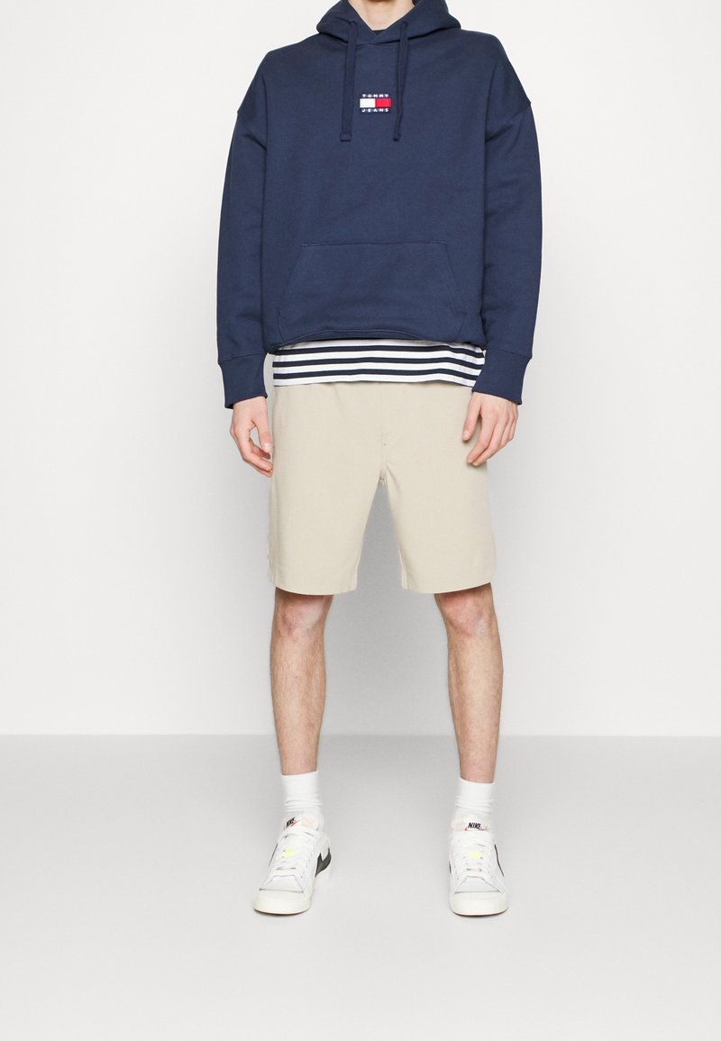 Personne portant un sweat à capuche Tommy Jeans bleu marine, un t-shirt à rayures en dessous, un short beige, des chaussettes blanches Nike et des baskets blanches, debout devant un fond blanc.
