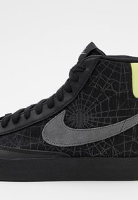 Nike high-top sneaker in black with textured fabric upper, gray suede accents, and a prominent gray swoosh logo. Features black laces and rubber sole.