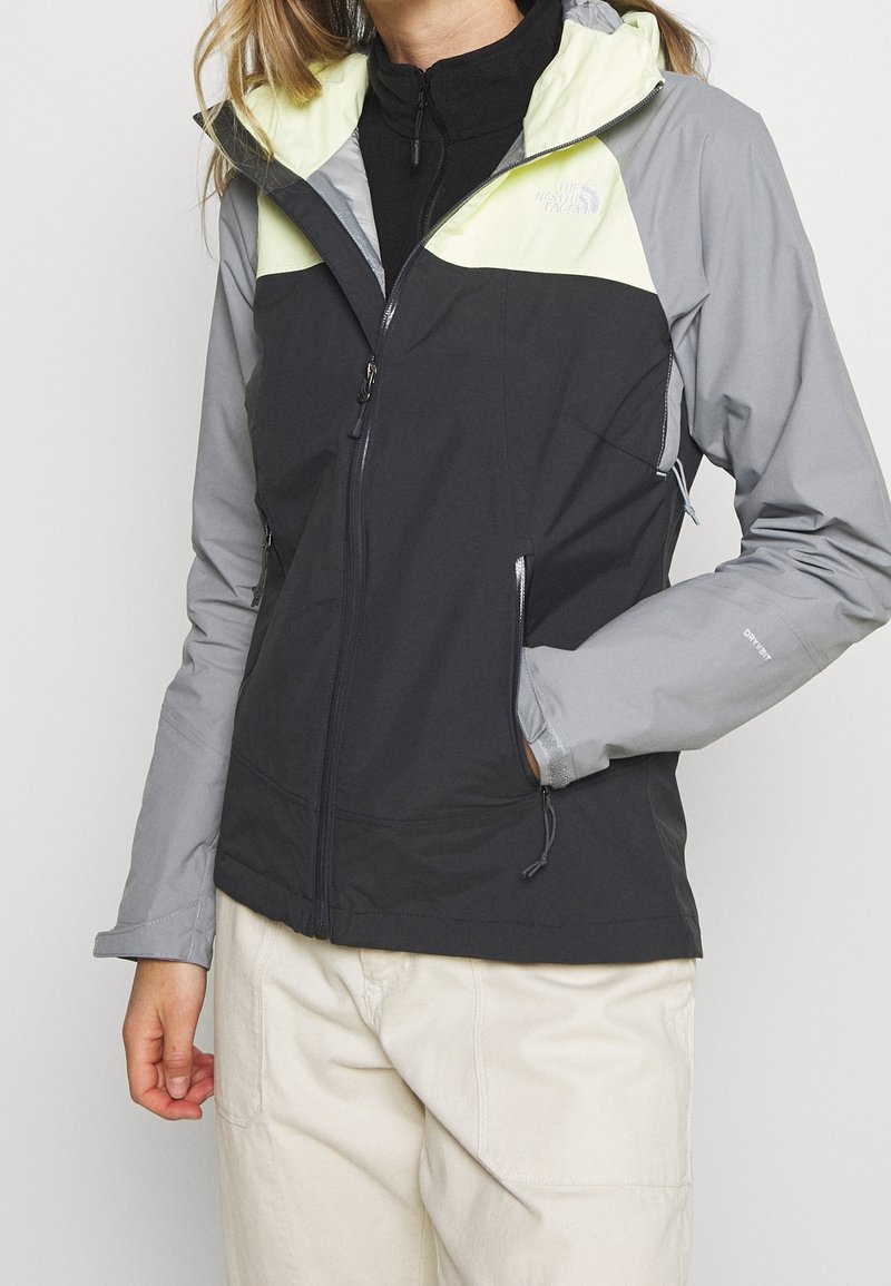 Femme portant une veste résistante aux intempéries noire, grise et jaune clair avec des poches zippées, un pantalon beige et un haut noir zippé en dessous.